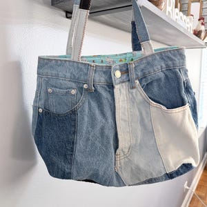 May include: A denim tote bag made from recycled jeans. The bag features a patchwork design with various shades of blue denim and white accents. It has two denim handles and a light blue patterned interior.