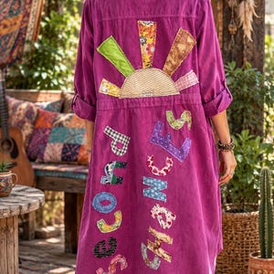 May include: A long-sleeved magenta button-down shirt dress featuring a patchwork sun design on the upper back and the words "second chances" appliqued in various patterned fabric letters across the lower back section of the garment.