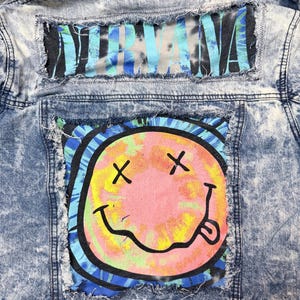 May include: Acid wash denim jacket with patches. The top patch reads "NIRVANA" in blue and gray lettering. The bottom patch features a smiley face with crossed-out eyes and a tongue sticking out, in pink, yellow, and black.