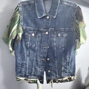Puede incluir: Un chaleco de mezclilla azul oscuro con cuello cl&aacute;sico, cierre frontal de botones met&aacute;licos y dos bolsillos en el pecho. La prenda est&aacute; decorada con mangas con volantes de tela de camuflaje verde y marr&oacute;n y un dobladillo a juego con lazos.