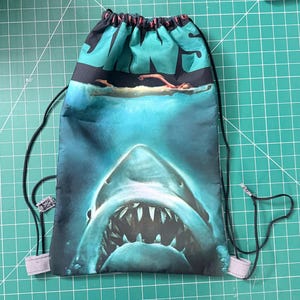 May include: A drawstring backpack featuring a movie poster design with the word "JAWS" in teal letters. The image depicts a swimmer in the water above a shark's open mouth. The bag has black drawstrings and gray fabric loops.