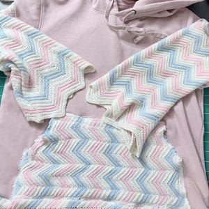 May include: A light pink hoodie with a matching crochet set. The crochet pieces feature a chevron pattern in white, light blue, and pink. The set includes a top and two sleeve pieces.