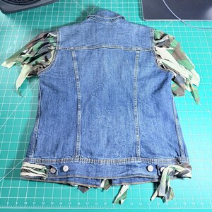 Puede incluir: Un chaleco de mezclilla azul con cuello cl&aacute;sico y cierre frontal de botones. Las sisas y el dobladillo inferior est&aacute;n adornados con tiras de tela de camuflaje verde, marr&oacute;n y negro, creando un efecto deshilachado y superpuesto en los bordes.