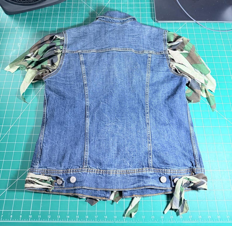 Puede incluir: Un chaleco de mezclilla azul con cuello cl&aacute;sico y cierre frontal de botones. Las sisas y el dobladillo inferior est&aacute;n adornados con tiras de tela de camuflaje verde, marr&oacute;n y negro, creando un efecto deshilachado y superpuesto en los bordes.