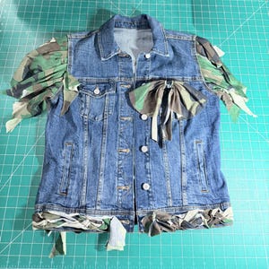 Puede incluir: Una chaqueta de mezclilla azul con mangas abullonadas de estampado de camuflaje, un gran lazo de camuflaje en el pecho y un borde de tela de camuflaje trenzado a lo largo del dobladillo inferior. La chaqueta incluye cuello est&aacute;ndar, botones de metal y dos bolsillos.