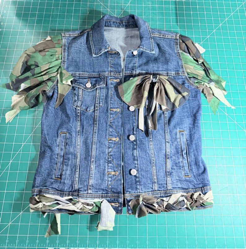 Puede incluir: Una chaqueta de mezclilla azul con mangas abullonadas de estampado de camuflaje, un gran lazo de camuflaje en el pecho y un borde de tela de camuflaje trenzado a lo largo del dobladillo inferior. La chaqueta incluye cuello est&aacute;ndar, botones de metal y dos bolsillos.