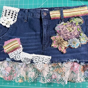 May include: Dark blue denim skirt with decorative patches and lace trim. Features floral embroidery and patches of lace and patterned fabric. The skirt has a ruffled lace hemline.
