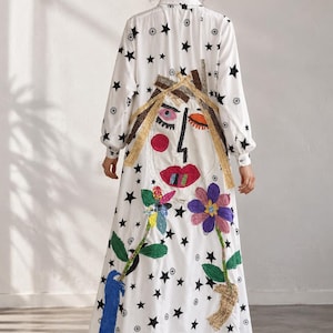 May include: A long-sleeved white maxi dress featuring a black star and sun pattern. The back displays a large, colorful fabric appliqué of a face with red lips, closed eyes, and textured hair, flanked by two floral stems with blue and purple petals.