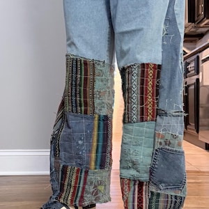 May include: Light blue denim jeans with patchwork detailing. The patchwork includes various patterned fabrics in shades of blue, red, and green. The jeans are paired with a white-soled shoe and a black boot.