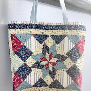 May include: A quilted tote bag featuring a central star pattern composed of navy blue, red, and cream fabric patches with floral and striped prints. The bag has two light blue fabric handles and a rectangular shape with visible stitching throughout.