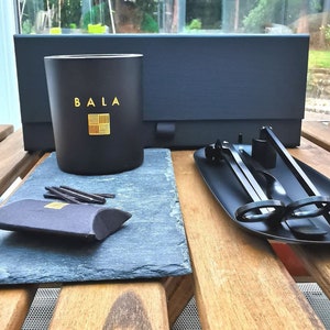 May include: A black candle jar with the word "BALA" in gold lettering on the front. The candle is on a black slate surface with a black box and a black metal candle snuffer on a tray.