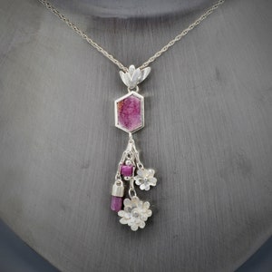 May include: A silver necklace with a pink hexagonal pendant and silver flower charms. The pendant is a faceted gemstone with a pink and white color.