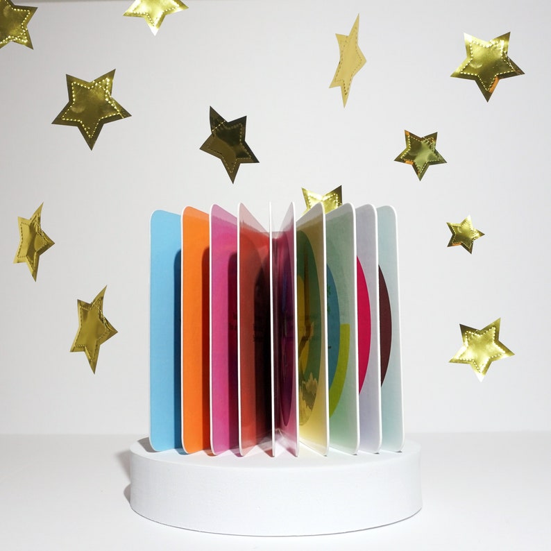 May include: A colorful pop-up book with a white base. The book has a rainbow of colors and is made of thick cardboard. The book is surrounded by gold stars.