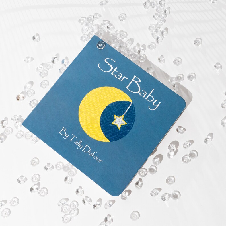May include: A blue book with the title "Star Baby" and the author "By Tally Dufour". The cover features an embroidered yellow crescent moon with a white star.