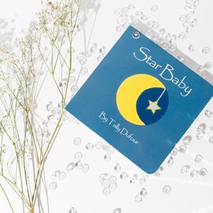 May include: A blue book with the title "Star Baby" by "Tally Dufour" and a yellow crescent moon with a white star on a blue background.