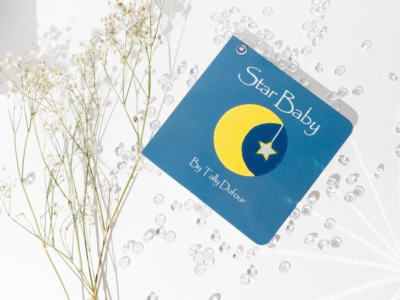 May include: A blue book with the title "Star Baby" by "Tally Dufour" and a yellow crescent moon with a white star on a blue background.
