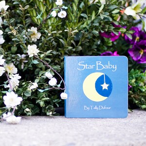 May include: A blue board book with the title "Star Baby" and the author "By Tally Dufour". The cover features a yellow crescent moon with a white star hanging from it.