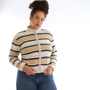 May include: A zip-up sweater with horizontal stripes in beige, navy, and yellow. The sweater has a high collar and is paired with blue jeans with distressed details.