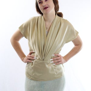 Könnte beinhalten: Eine goldfarbene Bluse mit kurzen Ärmeln und drapiertem Ausschnitt. Die Bluse hat eine taillierte Taille und wird mit einem hellgrünen Rock kombiniert. Das Model steht mit den Händen in den Hüften.