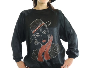 May include: Black long-sleeved sweatshirt featuring a graphic of a woman wearing a hat and gloves. The illustration is in shades of black, red, and gray. The sweatshirt has a ribbed hem and cuffs. The model is wearing a black hat and blue jeans.