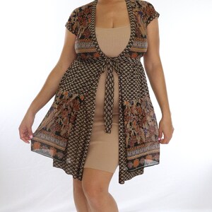 May include: A woman wearing a beige ribbed dress and a patterned, open-front kimono-style top. The kimono features a black background with floral and geometric designs in shades of brown, orange, and blue. She also wears beaded anklets.