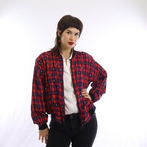 May include: A red and blue plaid bomber jacket with silver embellishments. The jacket has a black ribbed collar, cuffs, and hem. The jacket is worn over a white collared shirt and black jeans.