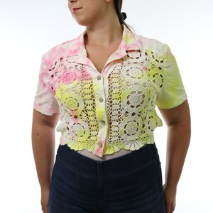 May include: A short-sleeved shirt with a tie-dye pattern in pink and yellow. The shirt features a crocheted floral design in yellow and white. The shirt is buttoned down the front and is paired with dark blue jeans.