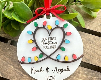 May include: White wooden Christmas ornament with a heart shape formed by a string of colorful lights. The ornament reads "Our First Christmas Together" and "Noah & Aryiah 2024".