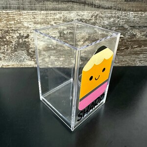 May include: A clear acrylic pencil holder with a yellow and pink pencil design and the word "SHARP" in black lettering.
