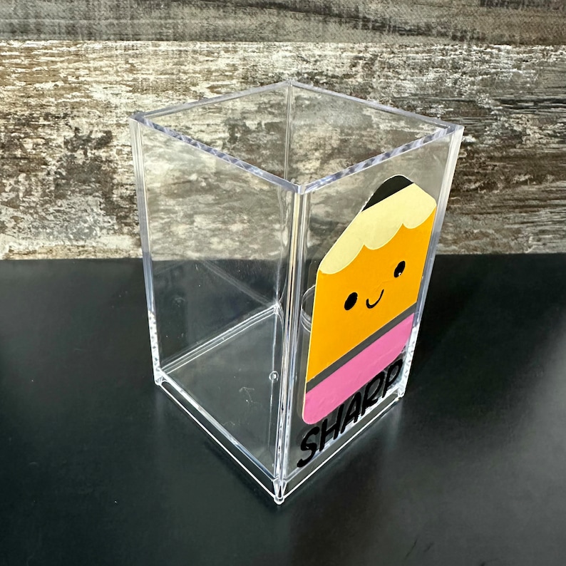 May include: A clear acrylic pencil holder with a yellow and pink pencil design and the word "SHARP" in black lettering.