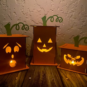May include: Three wooden jack-o'-lantern candle holders with different facial expressions. The candle holders are lit with small tea lights inside.
