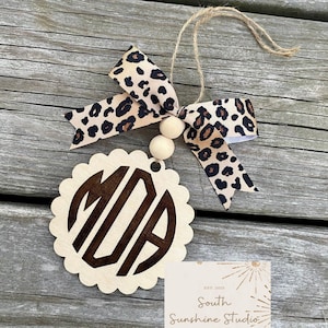 May include: A wooden monogram ornament with a scalloped edge, featuring the initials "MIA" in a dark brown font. It is adorned with a leopard print ribbon bow and wooden beads, suspended by a natural twine string. The ornament is from South Sunshine Studio.