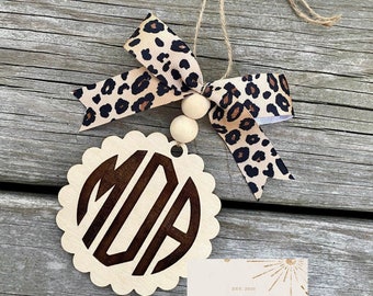 May include: A wooden monogram ornament with a scalloped edge, featuring the initials "MIA" in a dark brown font. It is adorned with a leopard print ribbon bow and wooden beads, suspended by a natural twine string. The ornament is from South Sunshine Studio.