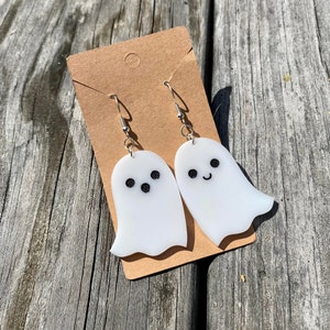 Op de afbeelding: Een paar witte oorringen in de vorm van een spook met zwart geverfde ogen en monden. De oorringen hangen aan zilveren haken op een bruine kaart.