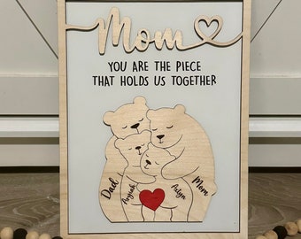 May include: Wooden sign with the text "Mom You are the piece that holds us together."  The sign features a cutout of a family of bears hugging each other. The bears are labeled "Dad", "Aryiah", "Aaliyah", and "Mom".