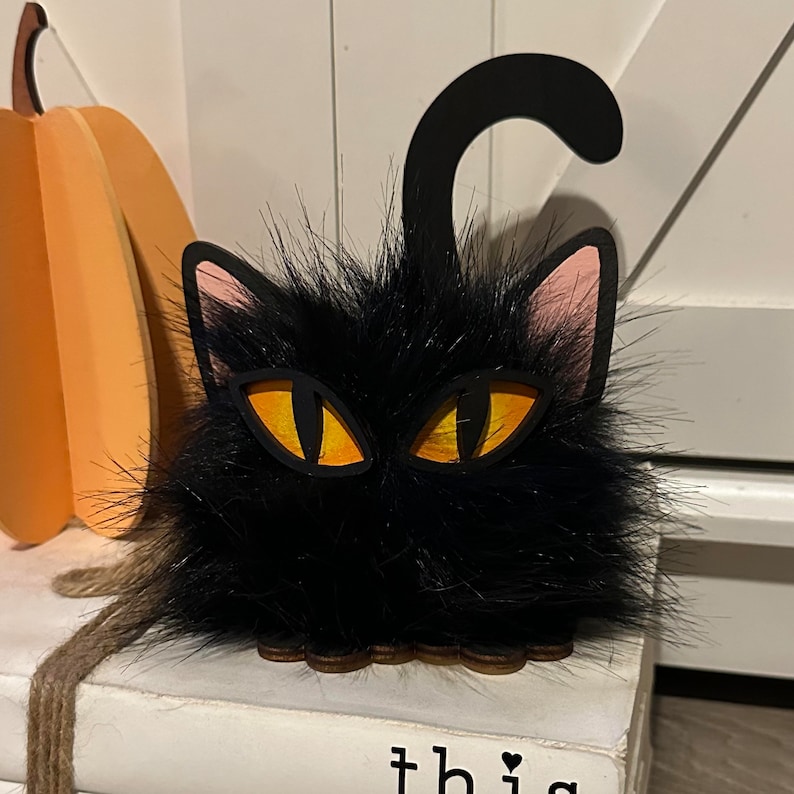 以下が含まれることがあります： オレンジ色の目とピンクの耳を持つ黒い猫のハロウィーンの飾り。猫は黒い毛皮でできており、木製の土台があります。
