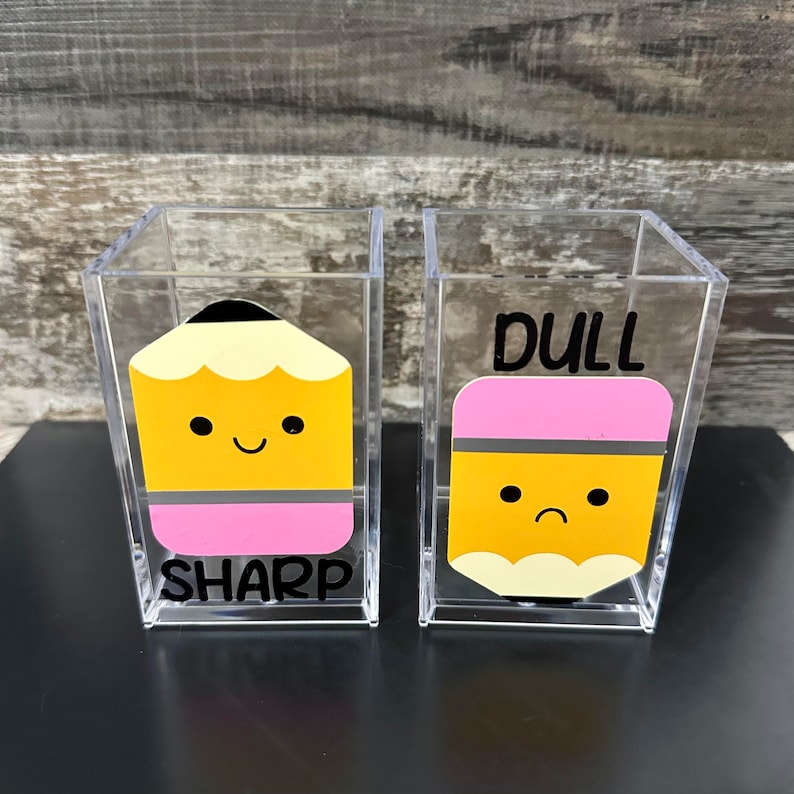 May include: Two clear acrylic pencil holders with cartoon pencils. The pencil on the left is smiling and says "SHARP". The pencil on the right is frowning and says "DULL".
