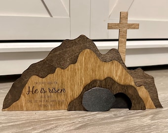 May include: Wooden Easter cross with a brown and gray mountain scene behind it. The mountain scene has a cave with a gray stone blocking the entrance. The text "He is risen" is engraved on the wood. The text "He is not here for as He said, Come, see the place where He lay" is also engraved on the wood. Matthew 28:6