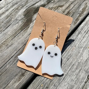 Op de afbeelding: Een paar witte oorringen in de vorm van een spook met zwarte ogen en een glimlachende mond. De oorringen zijn op een bruine kaart.