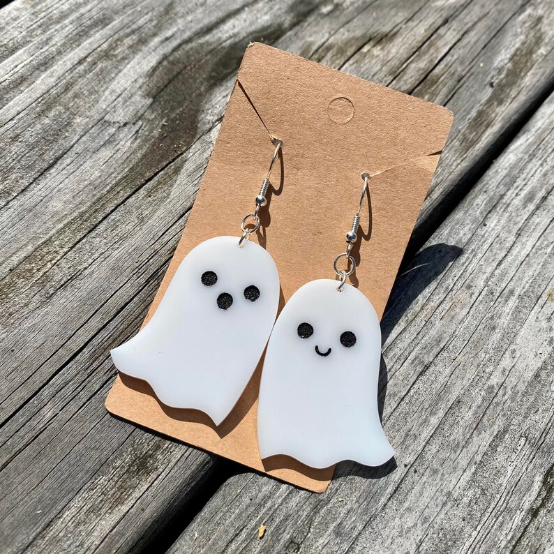 Op de afbeelding: Een paar witte oorringen in de vorm van een spook met zwarte ogen en een glimlachende mond. De oorringen zijn op een bruine kaart.