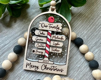 May include: A wooden Christmas ornament with a snow globe design. The ornament features a red and white candy cane with the words "Our Family" at the top. The names John, Gavin, Carter,  and  "Pussy" are on signs pointing to the left, and the names Melinda, Layla, and Fisher are on signs pointing to the right. The ornament also includes the year 2023 and the words "Merry Christmas".