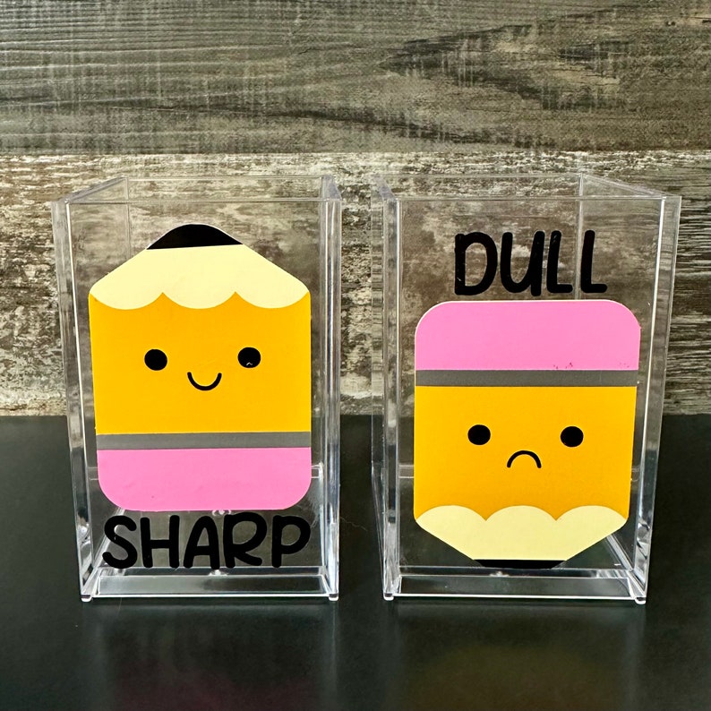 May include: Two clear acrylic pencil holders with cute cartoon pencils. One holder says "SHARP" and has a smiling pencil with a pink eraser. The other holder says "DULL" and has a frowning pencil with a pink eraser.