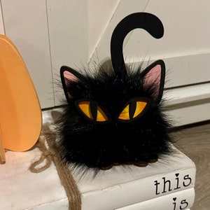 以下が含まれることがあります： ピンクの耳と黄色の目をした黒い猫の置物。猫は「this is」という文字が見える本の積み重ねの上に座っています。