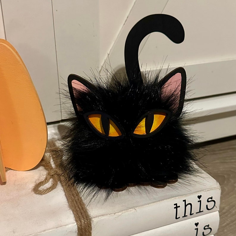 以下が含まれることがあります： ピンクの耳と黄色の目をした黒い猫の置物。猫は「this is」という文字が見える本の積み重ねの上に座っています。