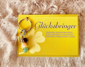 May include: A yellow card with the text "Glücksbringer" and a list of gemstones and their meanings. A silver key chain with a letter "H" charm and three gemstone beads is attached to the card.