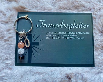May include: A silver key chain with a charm that has three beads, one orange, one white, and one black. The charm also has a silver letter "O" attached. The key chain is labeled "Trauerbegleiter" with the text "Sonnenstein | Hoffnung & Optimismus Bergkristall | Achtsamkeit Rauchquarz | Trauerbewältigung".