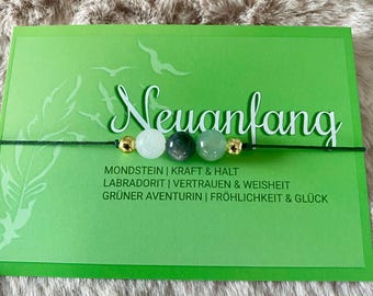 May include: A green card with the word "Neuanfang" written in white. The card has a black string with four beads: a gold bead, a white bead, a green bead, and a light green bead. The card also has text describing the meaning of each bead: "Mondstein | Kraft & Halt", "Labradorit | Vertrauen & Weisheit", "Grüner Aventurin | Fröhlichkeit & Glück".