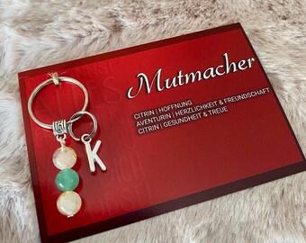 May include: A silver key chain with a charm that says "K" and three beads. The beads are green, white, and orange. The keychain is on a red card with the text "Mutmacher" and information about the stones.