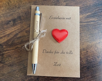 May include: A light-colored wooden pen engraved with the name "Emma" and a silver clip, tied with twine, rests on a brown card. The card features a red heart and German text.