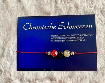 May include: Red, black, and gold beaded bracelet with a red string. The card reads "Chronische Schmerzen" and lists the stones and their properties: "ROTER JASPIS | löst ÄNGSTE & SCHMERZEN OBSIDIAN | löst VERSPANNUNGEN CITRIN | gegen KUMMER & STRESS"